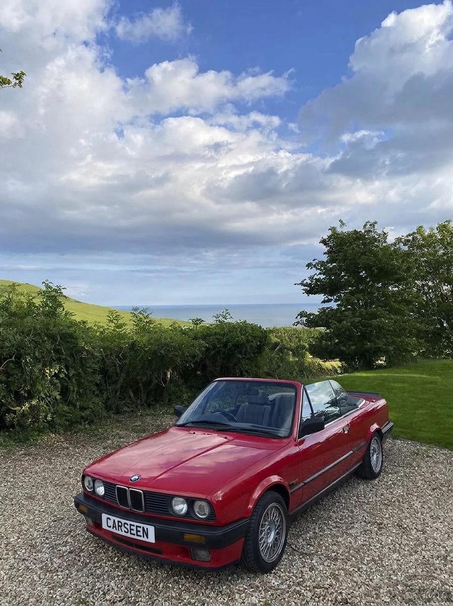 Red BMW E30 convertible on a gravel drive beneath a wide coastal sky