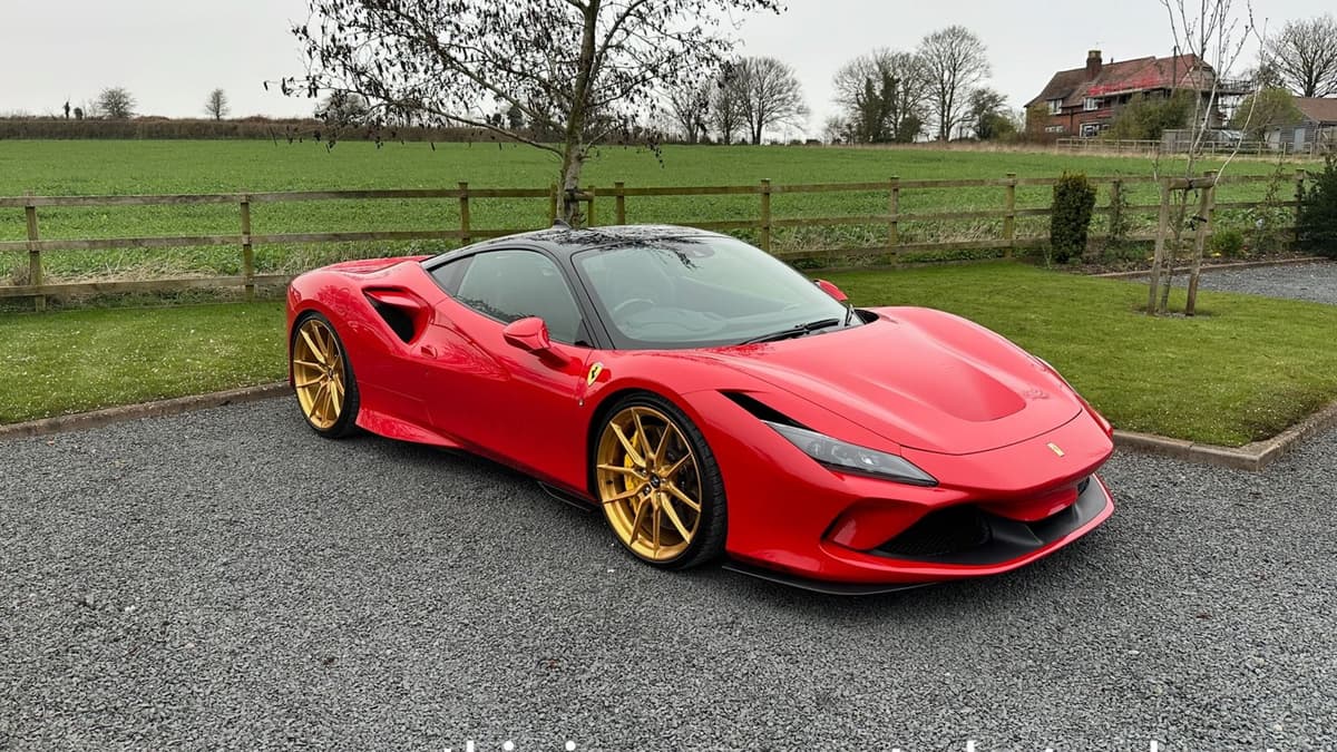 Red Ferrari F8 Tributo with gold wheels on a gravel drive beside green fields